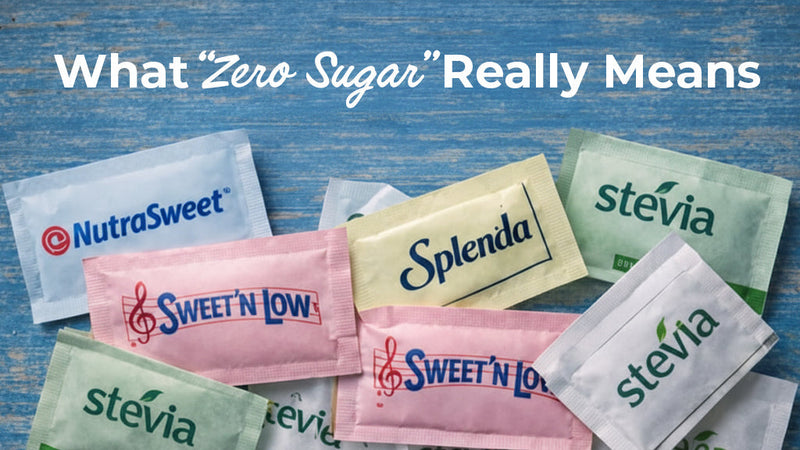 An overhead view of various artificial sweetener packets including brands like NutraSweet, Splenda, Sweet'N Low, and Stevia scattered on a blue wooden surface with text overlay reading "What 'Zero Sugar' Really Means."